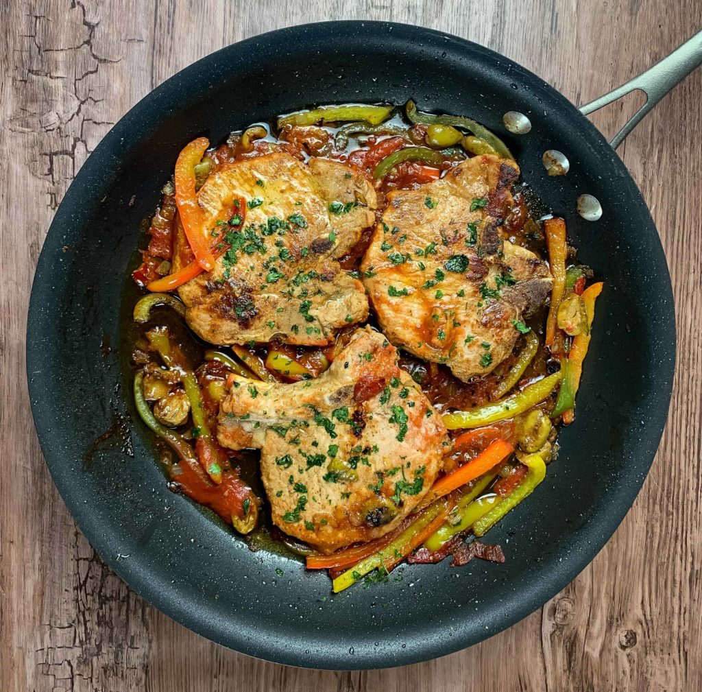 Braised Pork Chops in a skillet surrounded by peppers, olives, onions
