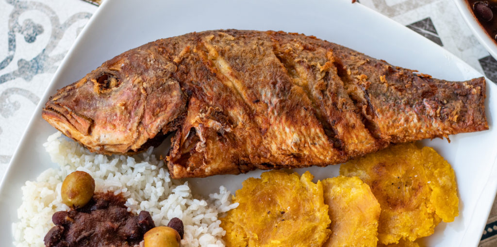 Fried red snapper whole on a plate with rice, beans and fried green plantains on the left
