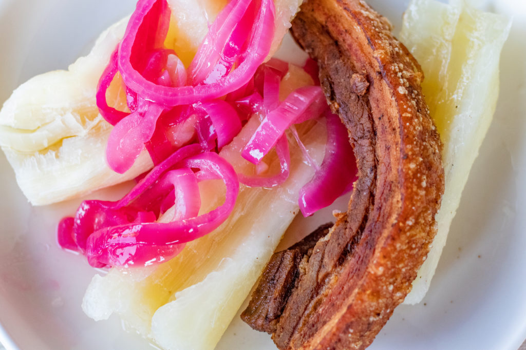1 dominican fried pork belly strip laying with boiled yuca and a red onion relish over the yuca