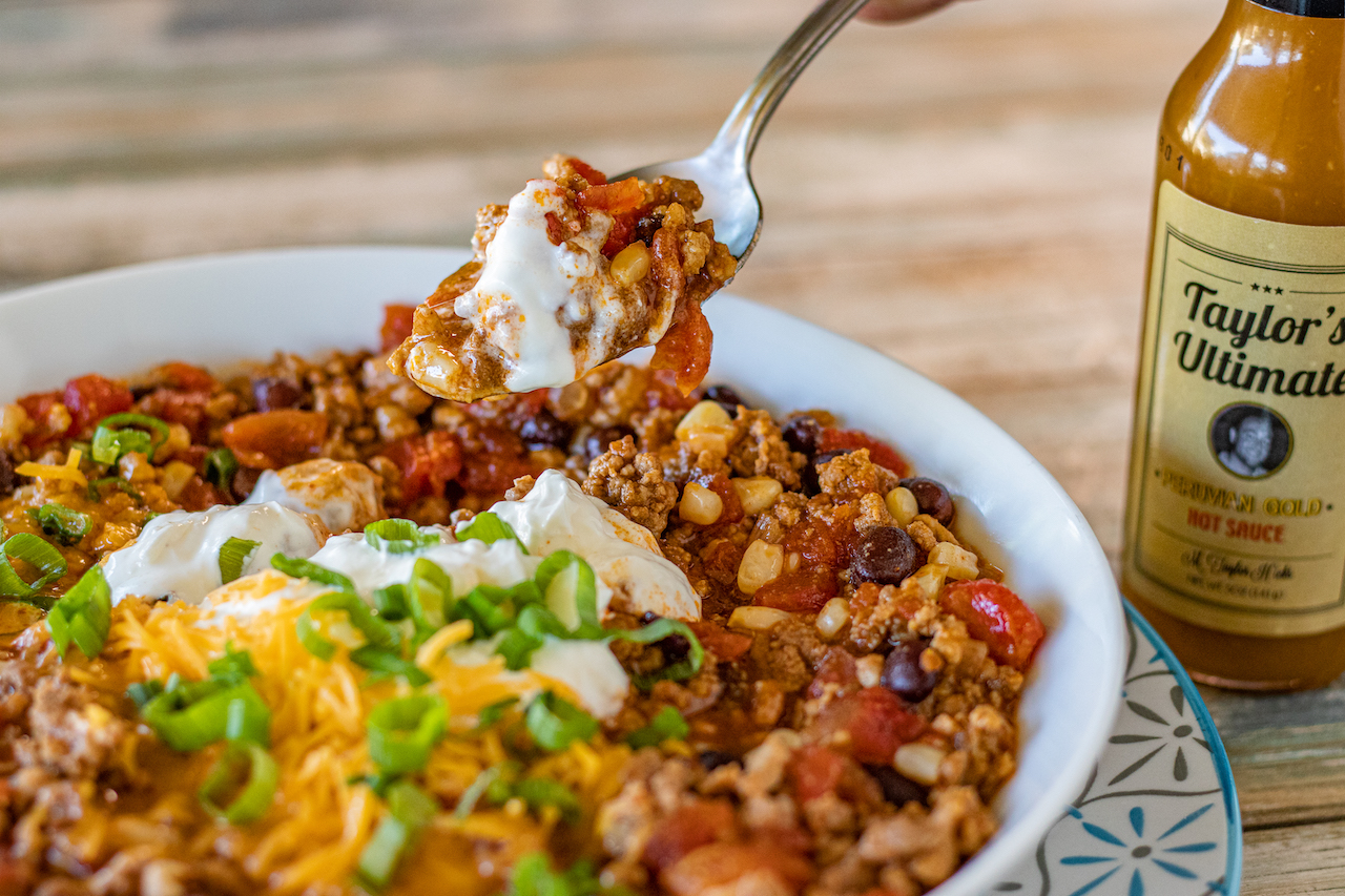 a shallow bowl of chili with shredded cheese, green onions and sour cream over it. A spoonful is being raised off the bowl.