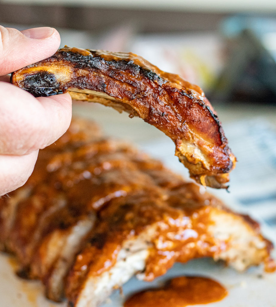 rack of cooked ribs in the background, blurred with a piece of the rib being held up to the camera