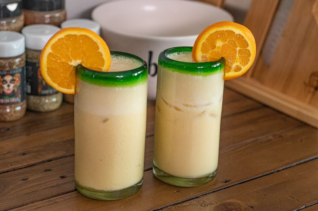 Dominican Morir Soñando - To Die Dreaming, a dominican republic traditional drink made with carnation milk, orange juice, sugar and vanilla extract. In this image you can see the finished drink in two tall glasses with an orange wedge for garnish sitting on the rims.