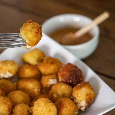 Goat Cheese Balls in a small serving platter. One is being held up towards the camera on a fork. You can see a tiny bowl in the back with honey and a honey dipper sitting in it.