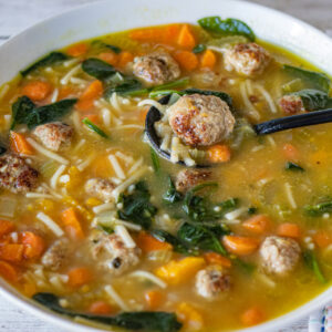A bowl of Italian wedding soup with a Latin Twist. There is a spoon holding one of the small meatballs in the recipe. You also see that this soup is made with fideos which is a cut spaghetti.