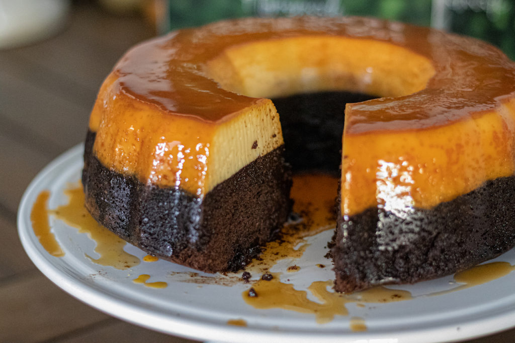 Flancocho, a mix. of flan and chocolate cake. Sitting on a cake stand with a view of it with a piece missing.