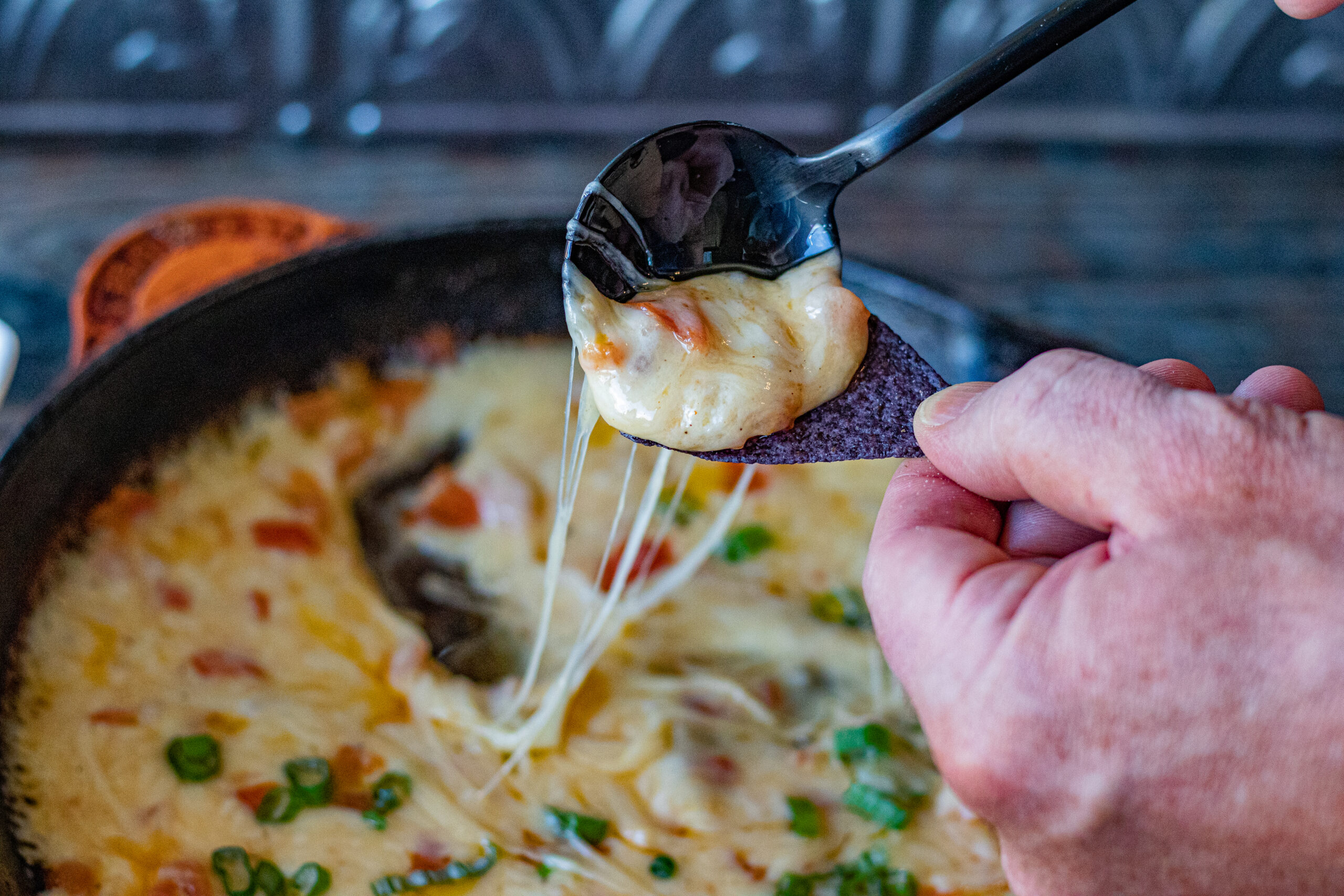 Mexican White Queso Dip in a cast iron skillet. A blue tortilla chip it being topped with it, with a spoon. You can see the cheese being super gooey and string as it is pulled to go on the chip.