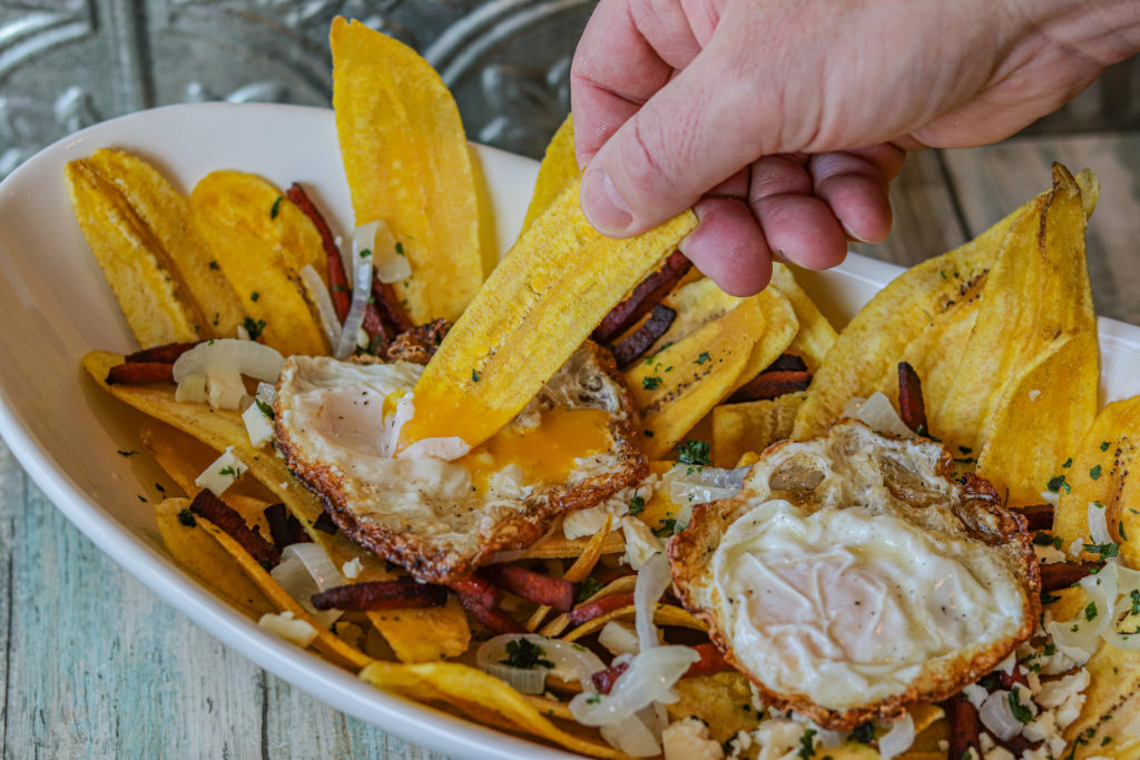 Loaded Dominican Plantain Chips on a serving platter. The chips are topped with fried salami strips, crumbled Queso Tropical, onion relish, fried eggs and chopped cilantro