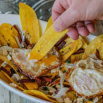 Loaded Dominican Plantain Chips on a serving platter. The chips are topped with fried salami strips, crumbled Queso Tropical, onion relish, fried eggs and chopped cilantro
