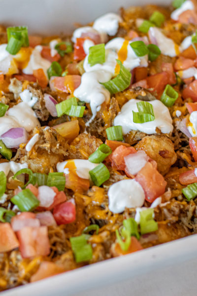 Loaded Pulled Pork Tater Tots in a baking dish. Topped with taco cheese, Mexican crema with lime juice, green onions and habañero honey mustard.