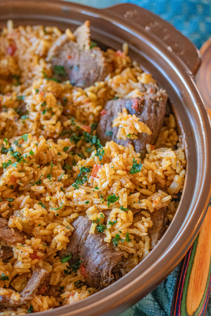 Image is of a One Pan Latin Beef and Rice in a serving dish. Here you only see half of the serving dish close-up. You can see the beautiful orange rice with thin slices of steak all around and topped with very finely chopped cilantro.
