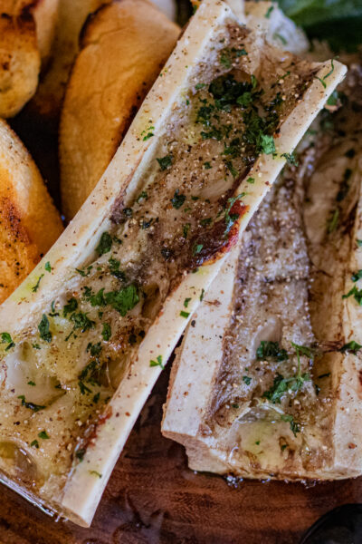 Roasted bone marrow simply seasoned with only a brushing of a good quality olive oil, sea salt and ground black and white peppercorn. In this image, you can see one bone marrow that has been sliced in half long ways. They are already roasted and accompanied by some sliced pan toasted french bread cut in slices. The bone marrow sits over top of mixed greens.