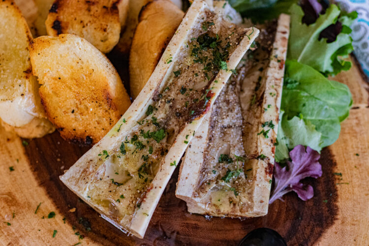Roasted bone marrow simply seasoned with only a brushing of a good quality olive oil, sea salt and ground black and white peppercorn. In this image, you can see one bone marrow that has been sliced in half long ways. They are already roasted and accompanied by some sliced pan toasted french bread cut in slices. The bone marrow sits over top of mixed greens.