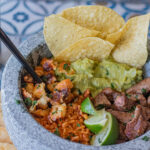 Quick Beef Tacos Molcajete Dinner - a Mexican molcajete filled with beef, yellow rice, fried panela cheese, guacamole, tortilla chips and lemon wedges. On the side you have corn tortillas and tostadas.