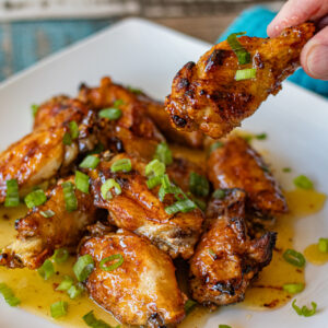 Sweet Chile Limon Chicken Wings on a plate. You can see the sweet honey mixture all over the wings and on the plate.They are topped with chopped green onions. One drumstick is being held up.