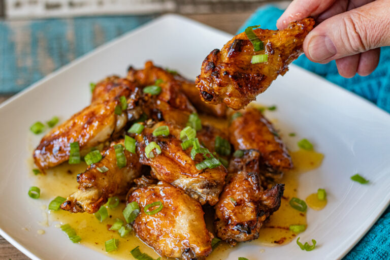 Sweet Chile Limon Chicken Wings on a plate. You can see the sweet honey mixture all over the wings and on the plate.They are topped with chopped green onions. One drumstick is being held up.