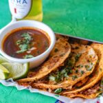 3 Instant Pot Beef Birria Tacos assembled on a serving platter with birria sauce on the side and lime wedges. A beer sit behind the platter.