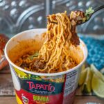 Instant Pot Beef Birria Ramen. Some of the noodles have the beef entangled and hover over the Ramen container.