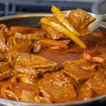 Dominican Carne Guisada in a skillet and a couple of pieces with peppers being held up showing you a closeup. Super delicious!