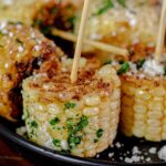 Elote (corn) Lollipops that are fried in vegetable oil for 4 minutes and seasoned with Spanglish Asadero's Elote Seasoning. In this image you can see several of them on a black plate. One full coirn on the cob cut in 4 round pieces. Once done, they are topped with the seasoning, chopped cilantro and crumbled Cotija cheese.