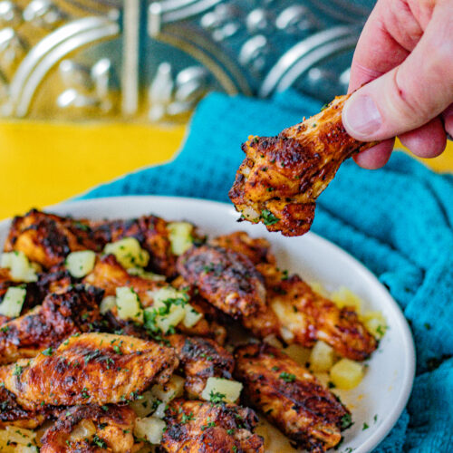 Al Pastor Chicken Wings on a serving plate and one drumstick being held to camera. You can see also the chopped up pineapple and chopped cilantro as garnishes in the background.