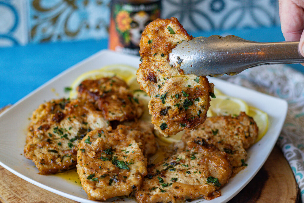 Lemon Carne Asada Pork Chops on a plate over lemon wedges. 8 pieces that are thin and boneless. One is being held up to show its deliciousness.