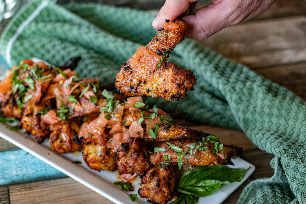 Chicken Wings with Tomato Dressing. A Spanish Tapas dish. O hand is holding one up. You can see bits of tomato and basil over the wings.