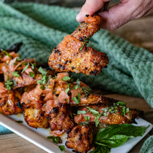 Chicken Wings with Tomato Dressing. A Spanish Tapas dish. O hand is holding one up. You can see bits of tomato and basil over the wings.
